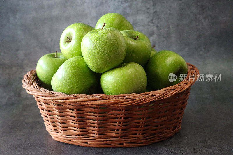 柳条篮子的特写图像，里面有一堆绿色的史密斯奶奶苹果(Malus domestica)，有茎，光滑的，有斑点的果皮，斑驳的深灰色背景，重点在前景